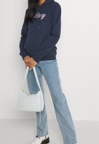 Sweat à capuche bleu marine avec texte brodé rose, assorti à un jean droit bleu clair et un sac à main vert à carreaux.