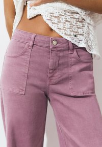 Mujer vistiendo pantalones de tiro alto rosas con grandes bolsillos frontales y una blusa de encaje blanca levantada para revelar el abdomen.