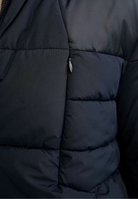 Section de doudoune en tissu bleu marine foncé, avec rembourrage matelassé, texture lisse, et une poche zippée sur le côté inférieur gauche.