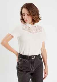 Weißes Kurzarmhemd mit Spitzenverzierung am Kragen, kombiniert mit schwarzen hochgeschnittenen Jeans und einem schwarzen Gürtel mit einer Metallschnalle.