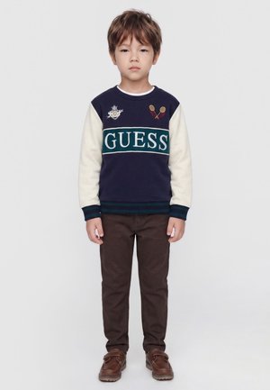 Jeune garçon debout droit portant un pull à manches longues bleu marine et blanc avec le logo "GUESS", pantalon marron et chaussures marron sur un fond uni.