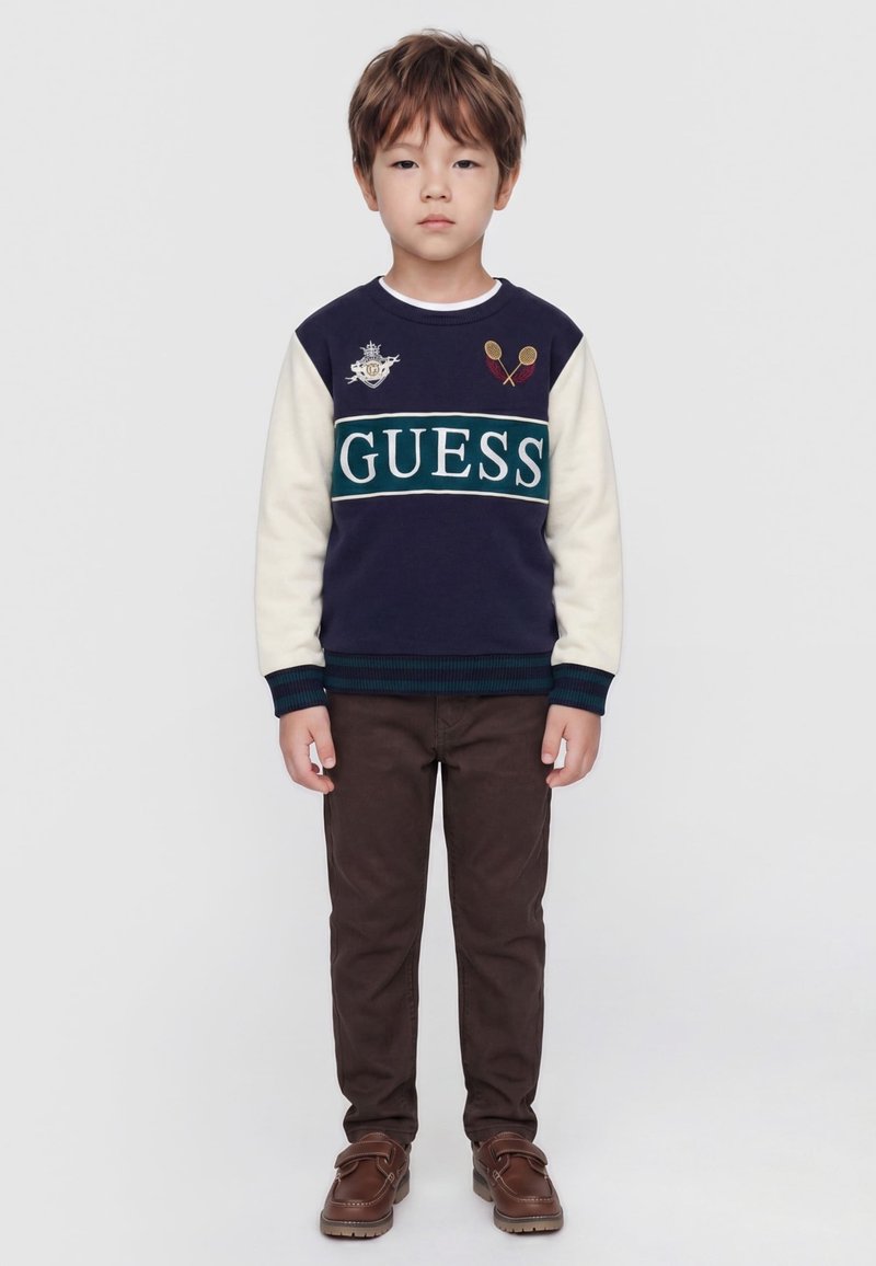 Jeune garçon debout droit portant un pull à manches longues bleu marine et blanc avec le logo "GUESS", pantalon marron et chaussures marron sur un fond uni.