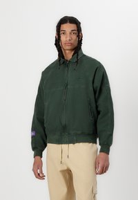 Grøn zip-up jakke med en afslappet pasform, der har en stående krave, sidelommer og en lilla labeldetalje på venstre ærme.