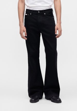Personne portant un jean évasé noir, des chaussures noires et une chemise blanche, debout devant un fond clair uni.