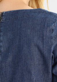 Prenda de mezclilla en azul oscuro, con una textura suave, escote redondo y cremallera visible en la parte posterior. El tejido es ligero y casual.