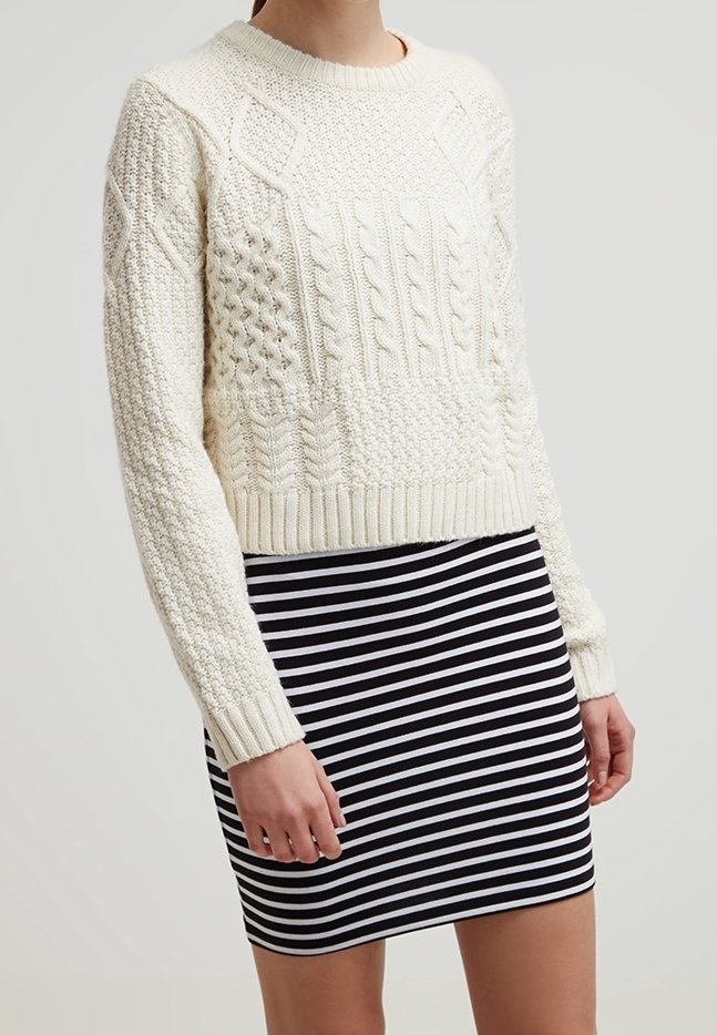 Personne portant un pull en maille blanche texturée et une jupe rayée horizontale noire et blanche debout devant un fond uni.