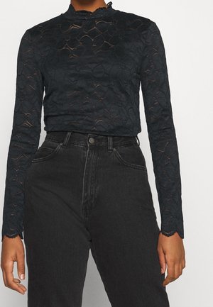 Haut en dentelle noire à manches longues avec un col haut, présentant un motif floral et des poignets en forme de scallop, associé à un jean foncé taille haute.