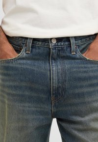 Os jeans de denim azul apresentam um corte reto, leve desgaste, detalhes em costura amarela e um clássico design de cinco bolsos com fecho de botão.