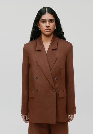 Femme aux longs cheveux noirs portant un blazer marron croisé à boutons noirs, debout sur un fond gris clair.