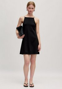Zwarte mouwloze mini-jurk met een rechte halslijn, gladde stof en een uitlopende zoom. Model houdt een zwarte clutch vast en draagt zwarte slippers.