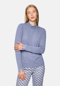 Langärmlige, hellilafarbene Bluse mit Kragen, die eine taillierte Passform und dekorativen Nähten aufweist. Kombiniert mit gemusterten blau-weißen Hosen.