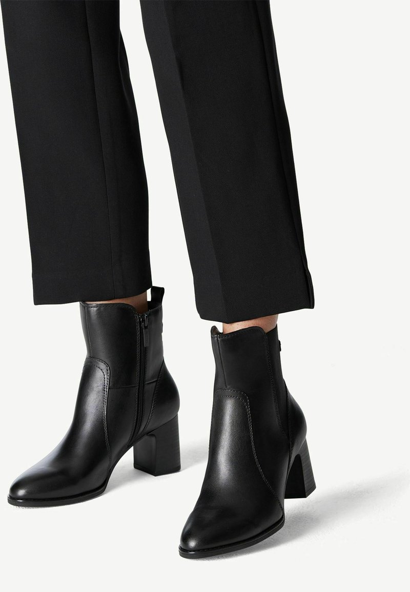 Bottes en cuir noires mi-mollet avec talons blocs et fermetures éclair latérales portées avec un pantalon noir court sur fond clair.