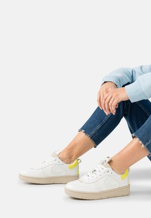 Baskets blanches avec une surface texturée, des accents crème et un talon jaune vif. Semelle beige clair et lacets plats complètent le design.
