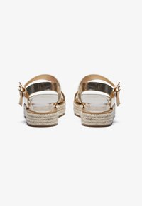 Gylne metalliske stroppesandaler med en spenne, med en espadrille-lignende vevd jute såle og flat hæl. Glatt tekstur.