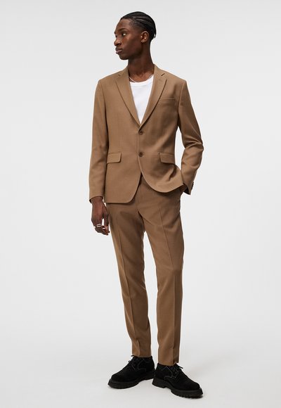 Brauner, maßgeschneiderter Anzug mit einem einreihigen Blazer, passenden Hosen und einem weißen Hemd. Schwarze Schuhe runden das Outfit ab.