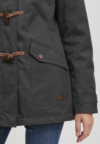 Grå ytterjakke med herringbone-mønster, med brune tau-lukkinger, en sidelomme og et lærlogo-merke.