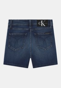 Calções de ganga em azul escuro com bolsos traseiros costurados e uma etiqueta de marca na cintura. Apresentam uma bainha reta e uma textura suave.
