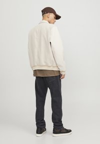 Vīrietis valkā beigu sporta jaku, brūnu cepuri, tumšas platās džinsus un melnus sporta apavus, stāv un skatās uz sāniem pret vienkāršu fona.