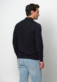 Mörkblå stickad tröja med långa ärmar och klassisk krage, tillverkad av ett mjukt tyg. Sedd från baksidan, tillsammans med ljusblå jeans.