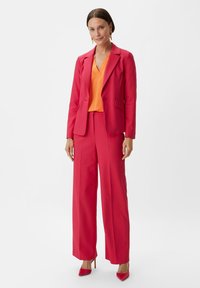 Heller rosa taillierter Blazer und weit geschnittene Hose, kombiniert mit einer orangen Bluse. Glatter Stoff, strukturiertes Design und spitze rote Absätze.