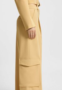 Beige op maat gemaakte broeken met een zachte textuur, wijdere pijpen en grote opgestikte zakken aan de zijkanten, met nauwkeurige stikdetails.