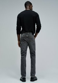 Suéter negro de manga larga combinado con jeans gris oscuro, con un corte clásico y bolsillos traseros, llevado con zapatos negros sobre un fondo neutro.