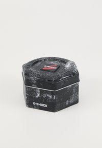 Kuusnurkne metallkonteiner musta värvi, tekstuursed pinnad. Kaanel on punane G-SHOCK logo ja aluse ümber hõbedane aktsent.