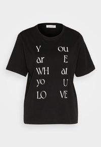 Camiseta negra de manga corta con texto blanco dispuesto verticalmente que dice "ERES LO QUE AMAS". Cuello redondo clásico, material de algodón.