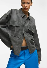 Szara skórzana koszula z kołnierzem, dwoma kieszeniami na piersi i luźnym krojem. W zestawie z jasnoniebieskimi spodniami z czarnym paskiem.