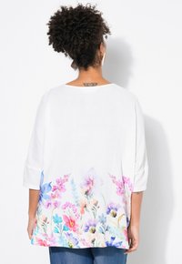 Tunic bianca con stampa floreale sul bordo in rosa, viola e blu. Presenta maniche a tre quarti e una vestibilità ampia. Tessuto testurizzato.