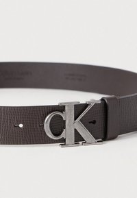 Καφέ ζώνη από δέρμα με υφή, με μια μεγάλη αγκράφα από ασημί μέταλλο σε σχήμα λογότυπου "CK". Ρυθμιζόμενο μέγεθος με πολλαπλές τρύπες για εφαρμογή.