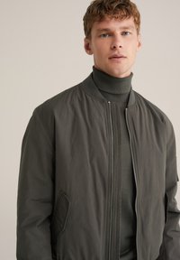 Mörkgrön bomberjacka med dragkedja fram, ribbad krage och sidofickor, lager på lager över en matchande grön polotröja.