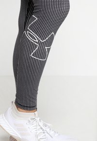 Mustat urheilulegginsit, joissa on ruutukuosi ja näyttävä valkoinen logo. Elastista materiaalia, joka istuu tiukasti, yhdistettynä valkoisiin urheilukenkiin.