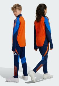 Jugend-Sportset mit orange-blauen Farbblöcken, langen Ärmeln und navyblauen Hosen mit orangefarbenen Streifen und Logo. Weiße Sportschuhe runden das Outfit ab.