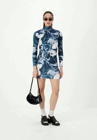 Γυναίκα φορώντας ένα μπλε και λευκό φλοράλ μακρυμάνικο φόρεμα, μαύρα γυαλιά ηλίου, μαύρη τσάντα χειρός, λευκές κάλτσες και μαύρες ανοιχτές σόλες παπούτσια.