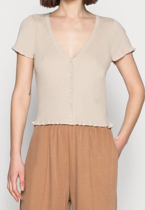 Kobieta ubrana w beżowy krótki, prążkowany top zapinany na guziki oraz brązowe spodnie z wysokim stanem, z jedną ręką w kieszeni.