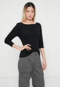 Jonge vrouw met steil ombre haar, gekleed in een zwarte top met lange mouwen en grijze cargobroek, poseert met haar hand op de heup tegen een witte achtergrond.