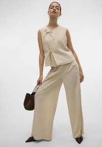 Vrouw poseert in een losse beige mouwloze top en broek met brede pijpen, met een donkerbruine handtas en puntige bruine schoenen.