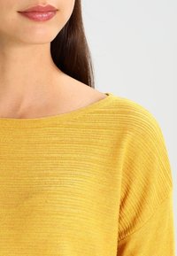 Der langärmlige gelbe Pullover zeichnet sich durch ein weiches, strukturiertes Strickmuster mit einem weiten Bootsausschnitt und einer lockeren Passform aus, die eine lebendige, einfarbige Optik zur Geltung bringt.