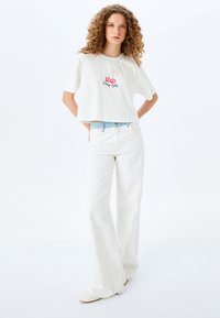 Weißes, kurzes T-Shirt mit Kirschgrafik und dem Text "Cherry Baby", kombiniert mit hoch taillierten, weißen weit geschnittenen Jeans und cremefarbenen Slippern.