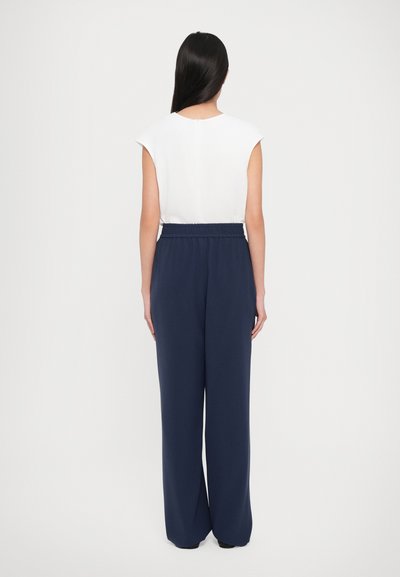 Woman standing facing away, wearing a sleeveless white top and high-waisted, wide-leg navy blue trousers on a plain background.