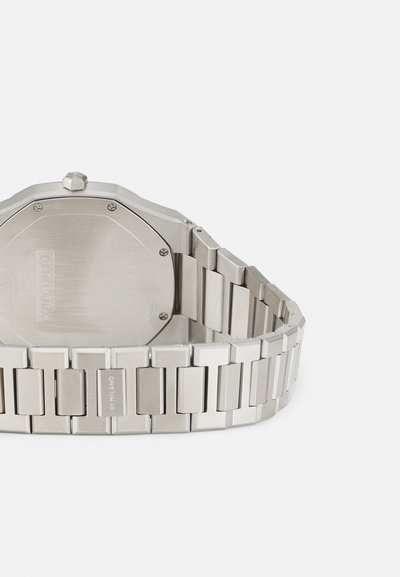 Stříbrné nerezové hodinky s osmibokým ciferníkem a řetízkovým náramkem v obdélníkovém tvaru. Mají lesklý povrch a vyryté jméno značky na pásku.