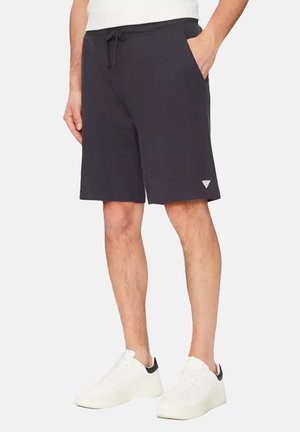 Homme portant un short noir à cordon avec poches latérales et des baskets blanches avec des languettes noires au talon.