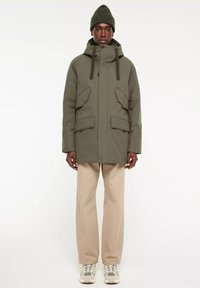 Olijfgroene parka jas met een capuchon, voorzien van grote voorzakken, een knoopsluiting en een losse pasvorm, gecombineerd met beige broek en sneakers.