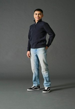 Marinefarbener Reißverschluss-Pullover, leicht ausgebleichte Jeans und schwarze Sneakers. Das Modell steht lässig mit einer Hand in der Tasche vor einem grauen Hintergrund.