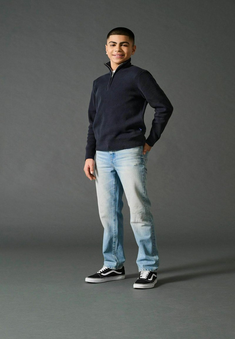 Jūras zila sporta jakas, gaiši nodilušas džinsus un melnas kedas. Modelis stāv brīvā pozā, ar vienu roku kabatā uz pelēka fona.