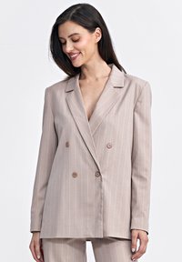 Dwurzędowa marynarka w jasnym beżowym kolorze z białymi prążkami. Posiada klapę i brązowe guziki z przodu.