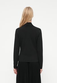 Blonda sieviete ar taisniem matiem valkā melnu garām piedurknēm topu un kroklu, stāvot ar muguru pret baltu fonu.