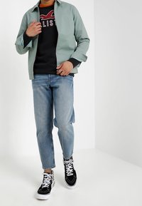 Light green jacket over a black sweatshirt with red lettering, paired with blue jeans and black and white patterned sneakers.