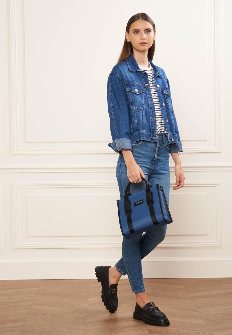 Sac à main à poignées bleues avec des accents noirs, au design structuré, associé à une veste en denim, une chemise rayée et un jean. Texture lisse, tenue décontractée.
